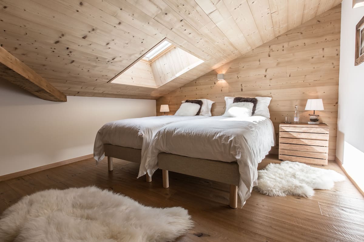 A bedroom with a sloped ceiling, a skylight, a bed with white bedding, a wooden nightstand with a lamp, and a fluffy rug on the floor.