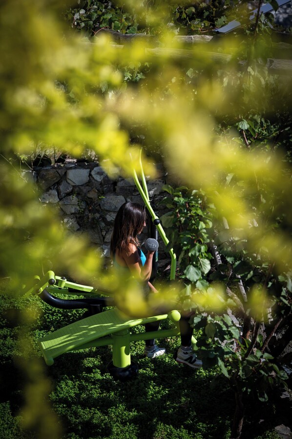 Una persona sentada en un banco verde para ejercicios en un jardín, con un muro de piedra y follaje al fondo.