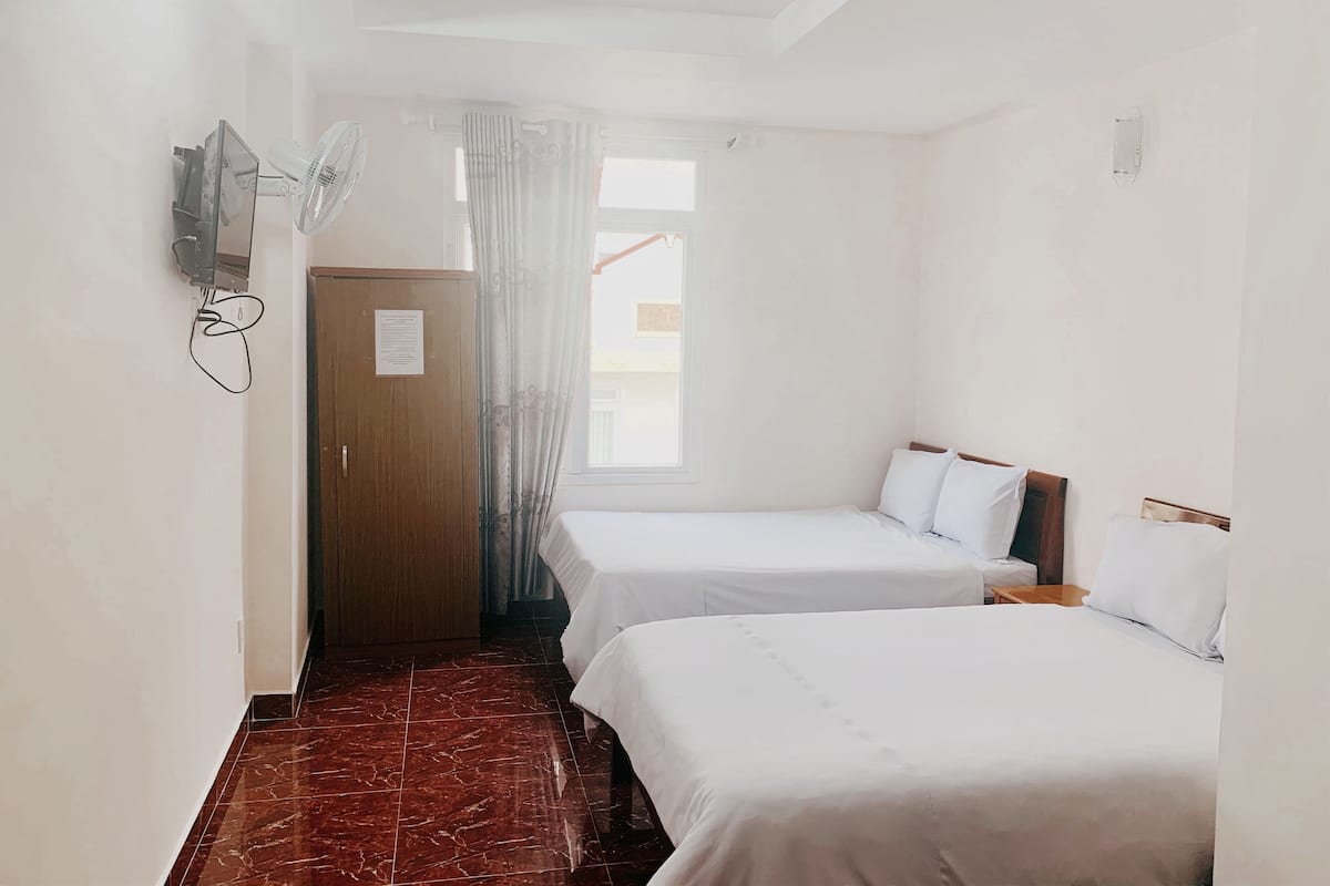 A hotel room with two beds, a wooden wardrobe, a wall-mounted TV, a window with curtains, and a tiled floor.