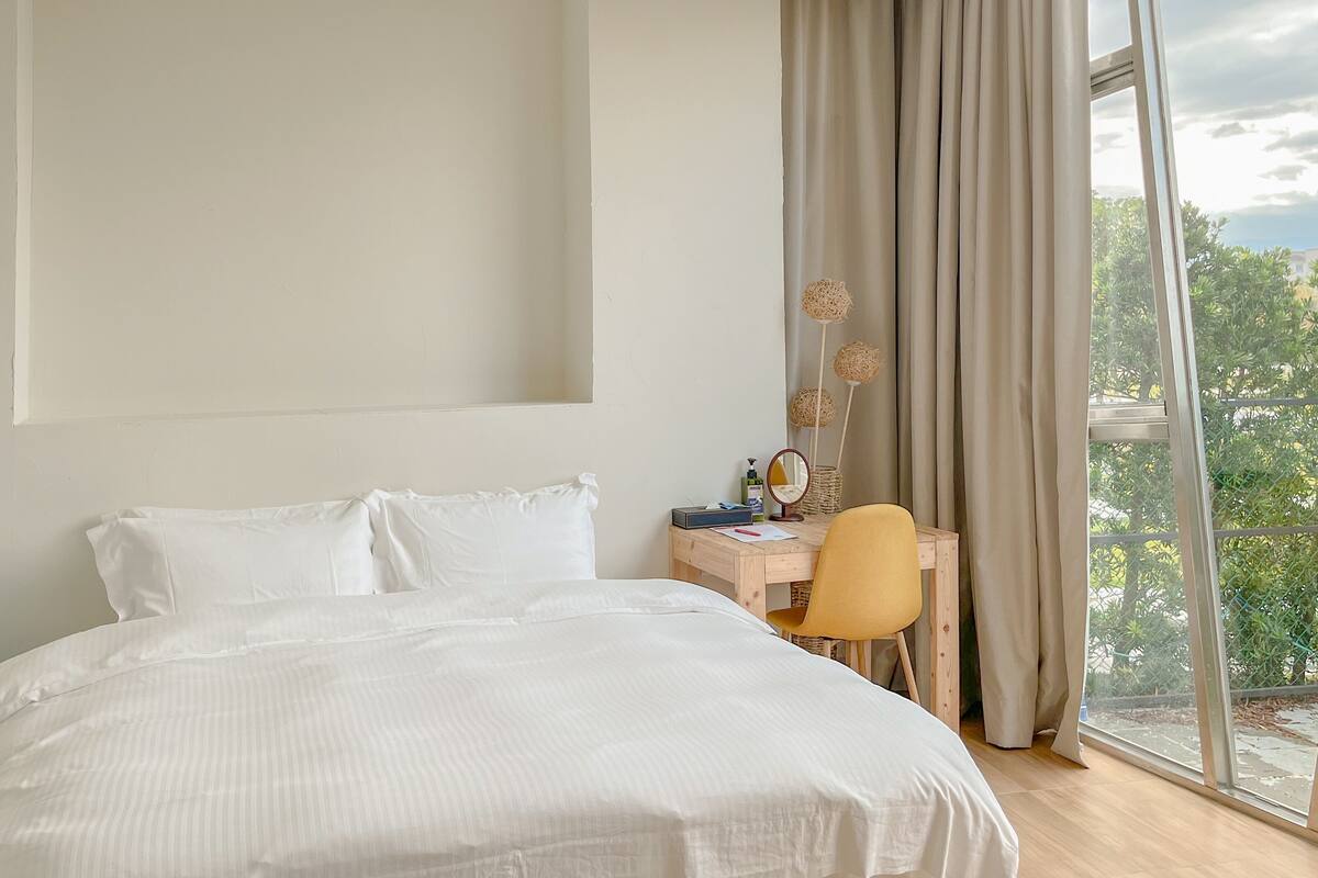 A hotel room with a large bed, a desk with a chair, a window with curtains, and a view of greenery outside.