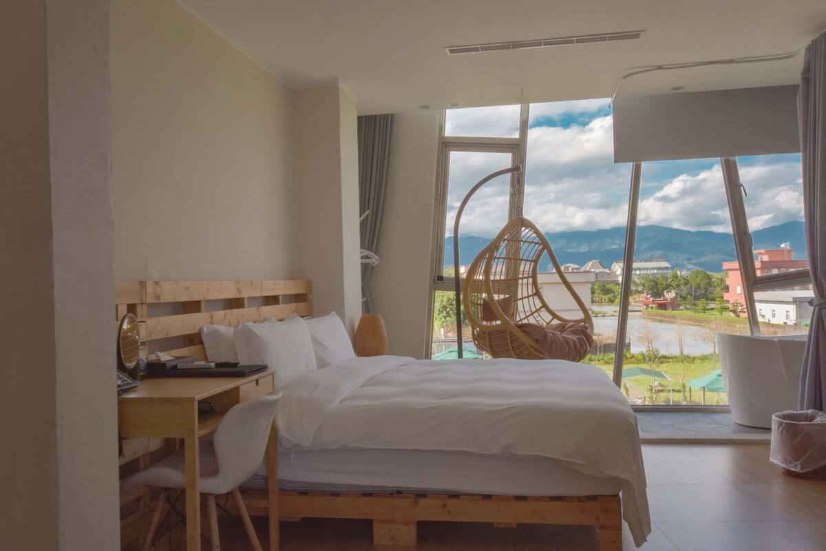 A bedroom with a wooden bed, a desk, and a chair. There is a large window with a view of mountains and a body of water.