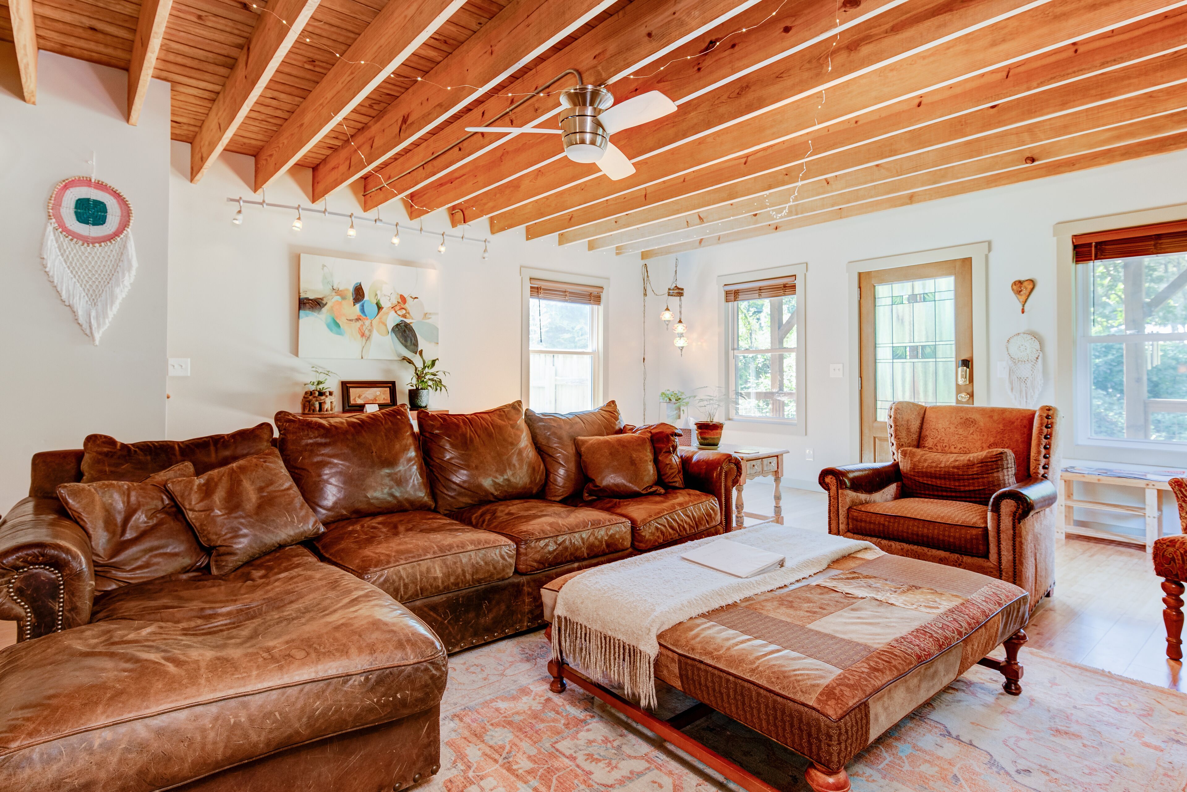 A spacious living room with a wooden ceiling, leather sofas, a wooden coffee table, and a large window providing natural light.