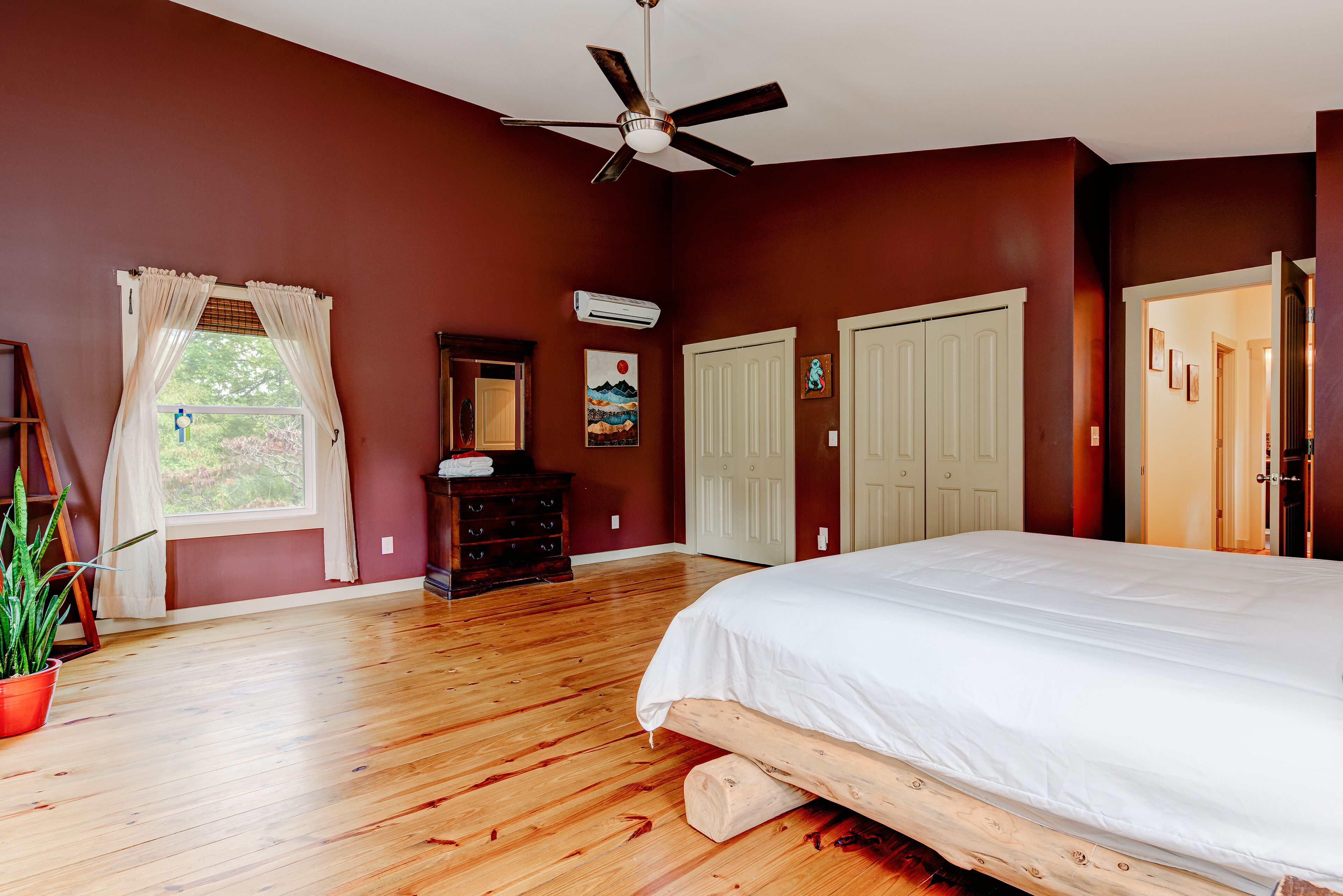 A bedroom with a wooden floor, a bed, a ceiling fan, a dresser, a window with curtains, and a door.