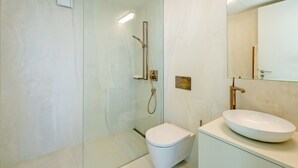 Egy modern fürőszoba, üvegfalas zuhanyzóval, fehér WC-vel és egy nagy, ovális mosdókagylóval, melynek felett tükör található.