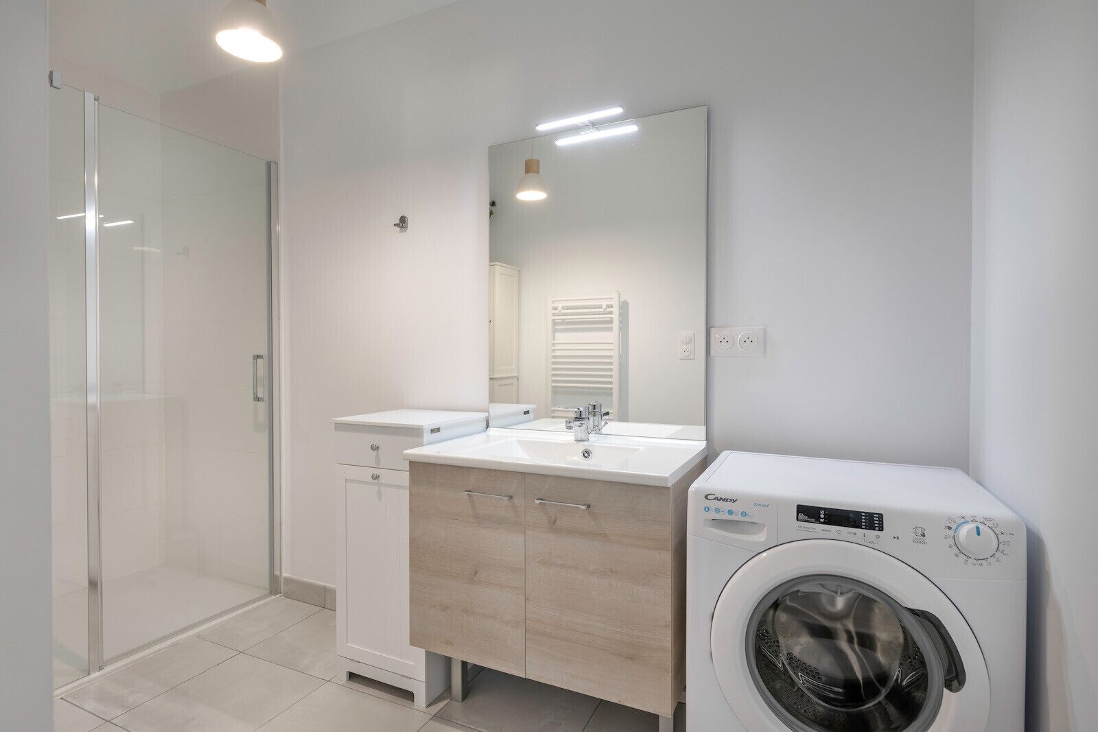 Ein modernes Badezimmer mit Duschkabine aus Glas, einem weißen Waschbecken mit Spiegel darüber und einer Waschmaschine mit Frontlader.