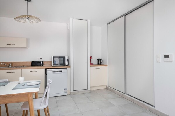 Una cocina compacta con microondas, tostadora y cafetera sobre la encimera, lavavajillas y comedor con mesa y sillas.