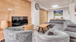 A modern living room with a flat-screen TV, grey sofas, a wooden accent wall, a clock, and a painting of a landscape. - Val Louron, 15-pers. apartment near slopes (Genos)