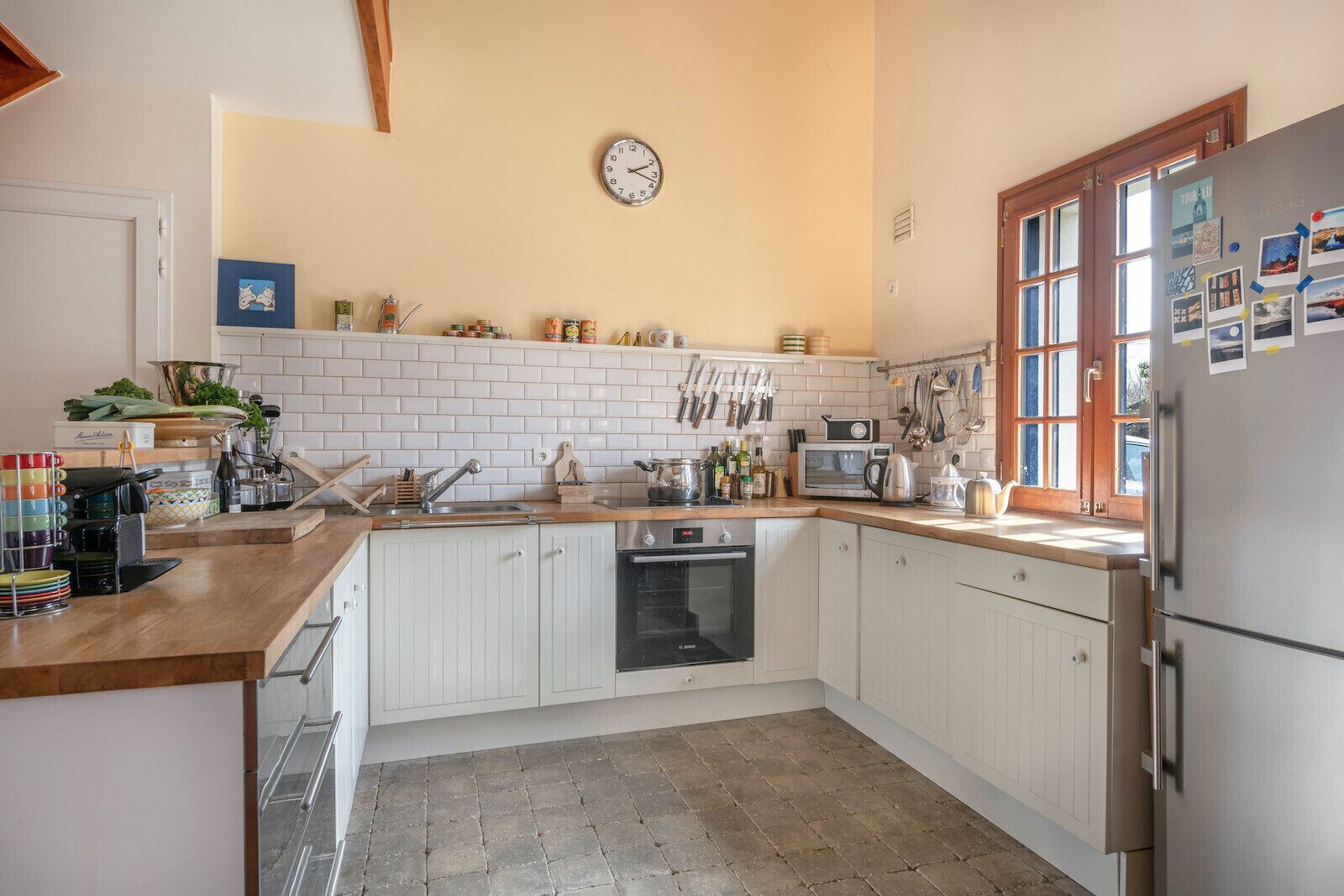 A kitchen with white cabinets, a microwave, a sink, and a refrigerator. There is a clock on the wall and a window with multiple panes.