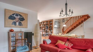 Una sala de estar con un sofá rojo, estanterías de madera, un sillón de madera, una mesa de centro de vidrio, piso de madera y una escalera.
