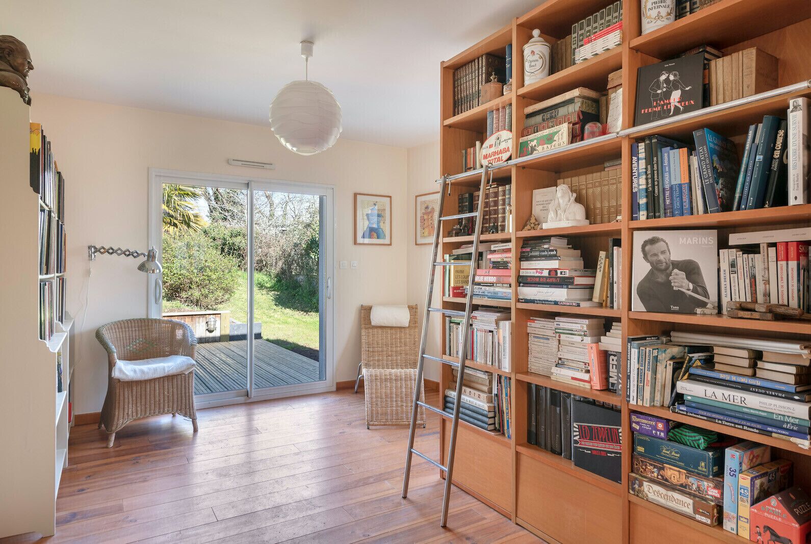 Une pièce avec une grande bibliothèque, un fauteuil en osier, une échelle et une vue sur l’extérieur, visible à travers une porte vitrée.
