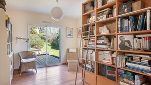 Una habitación con una amplia estantería, un sillón de mimbre, una escalera y vistas al exterior a través de un ventanal.