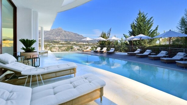 Un área de piscina exterior moderna con sillones de descanso y sombrillas, con vistas a un paisaje montañoso.