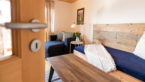 Une pièce chaleureuse avec un plafond en bois, un lit doté d’un matelas bleu, une table en bois, une petite table d’appoint avec une lampe et une fenêtre avec des rideaux.