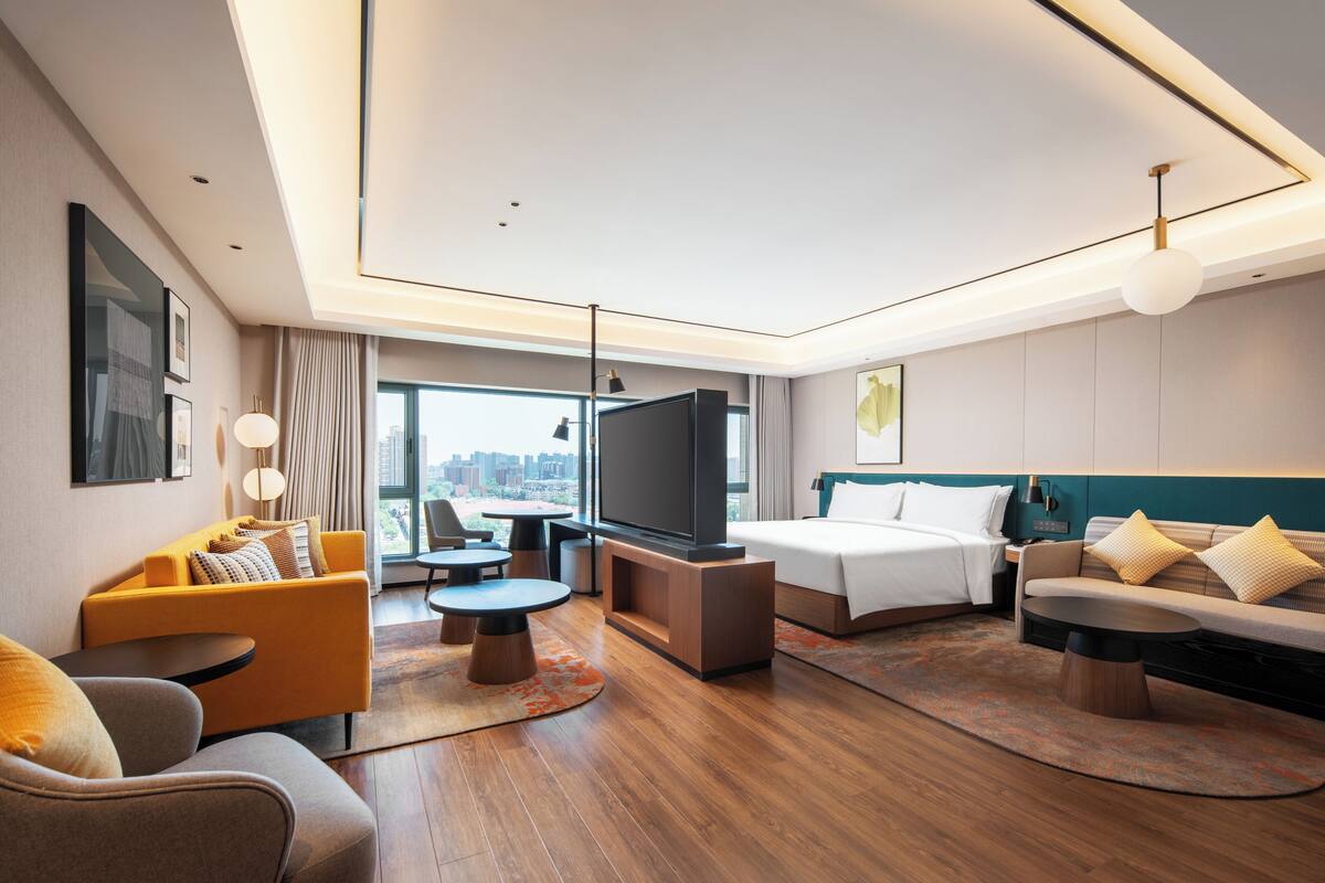 A modern hotel room with a bed, sofa, TV, and city view.