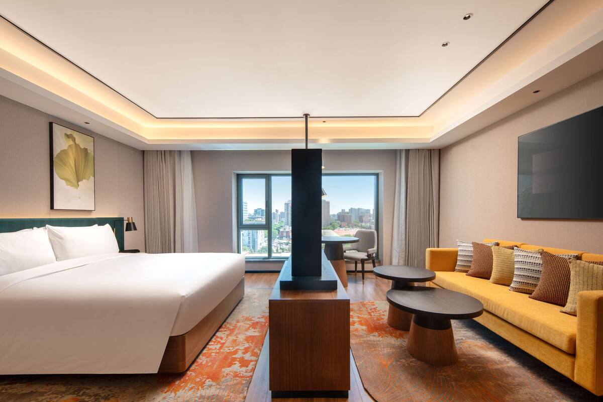 A modern hotel room with a large bed, a sofa, a dining table, and a city view through the windows.