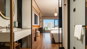 A modern hotel room with a large bed, a desk, a bathroom with a mirror, and a view of the city.