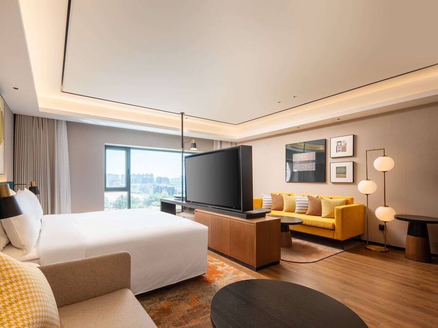 Ein modernes Hotelzimmer mit einem großen Bett, einer Couch, einem Fernseher und Blick auf die Stadt.