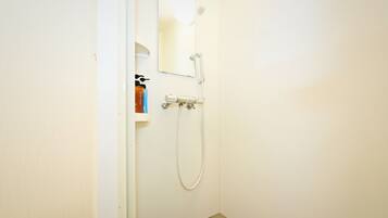 A small, white bathroom with a shower, a mirror, and a shelf with toiletries.