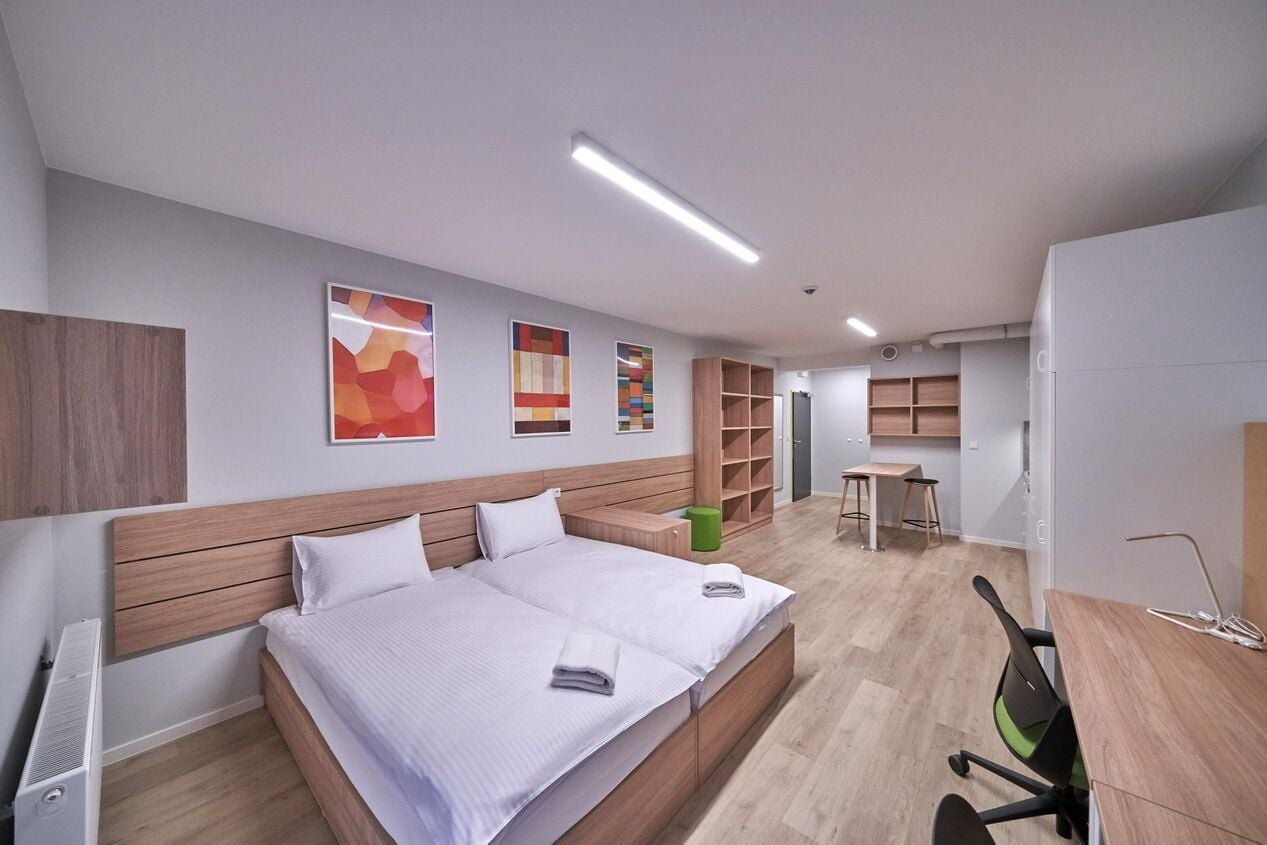 Een moderne hotelkamer met een groot bed, een bureau en een stoel. Er zijn planken en een prullenbak in de kamer.