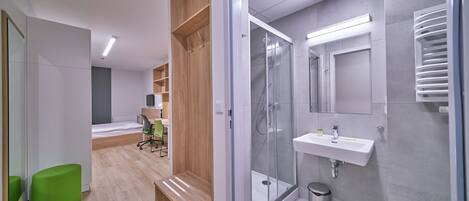 Une chambre d’hôtel moderne avec un bureau en bois, une chaise verte et un espace douche doté d’une porte en verre.