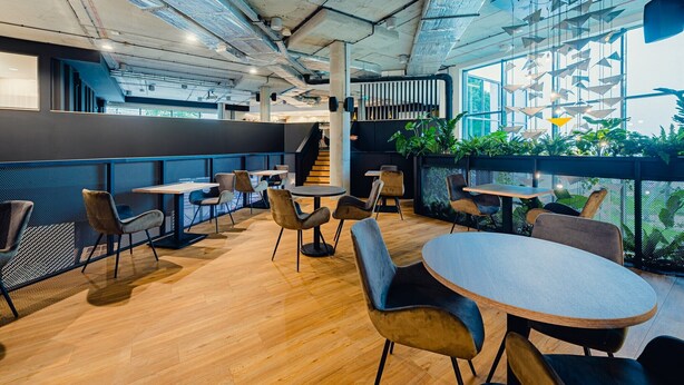 A modern indoor space with wooden flooring, round tables, and chairs. There are large windows allowing natural light and views of greenery outside.
