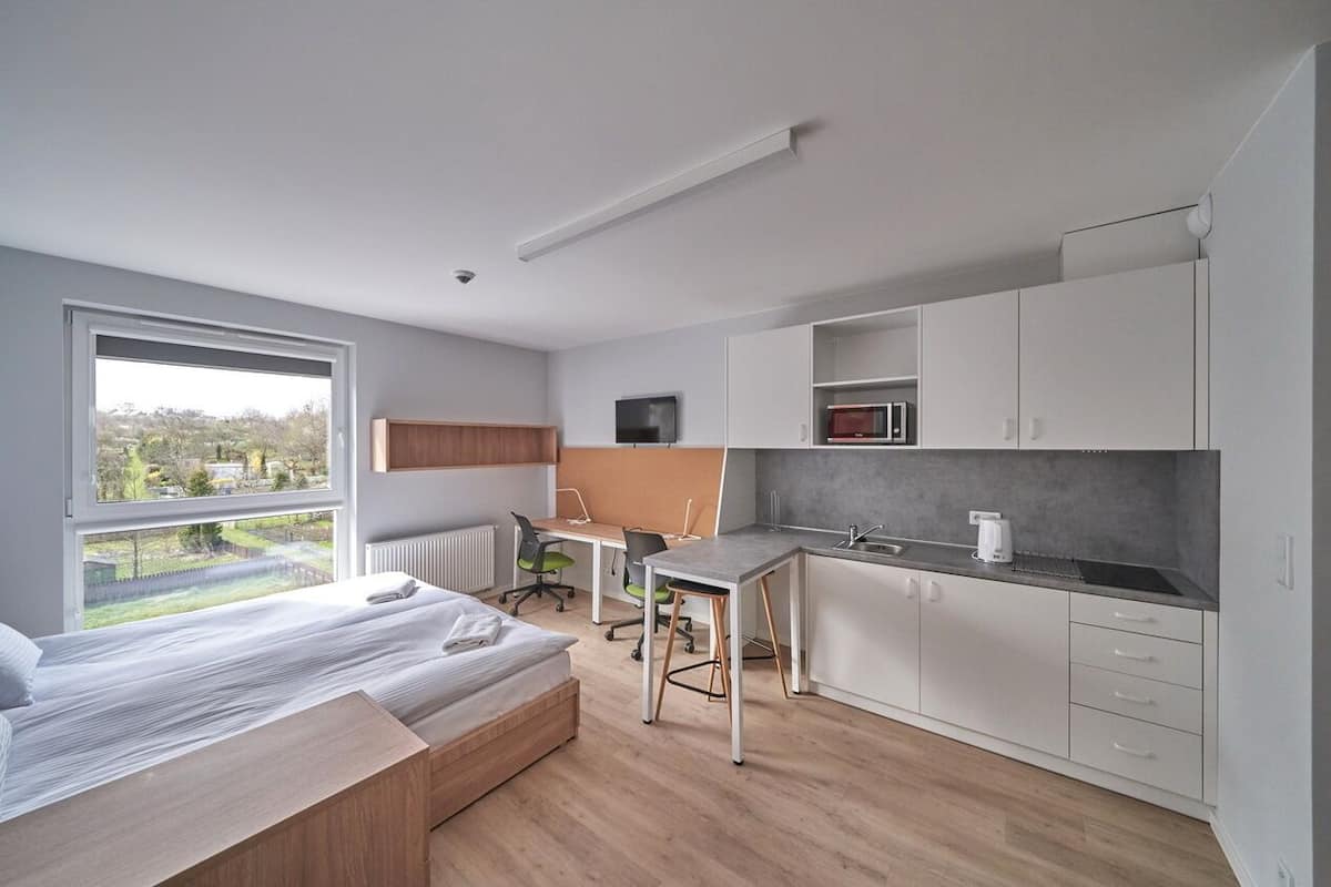 A modern bedroom with a large window, a bed, a desk, and a kitchenette.