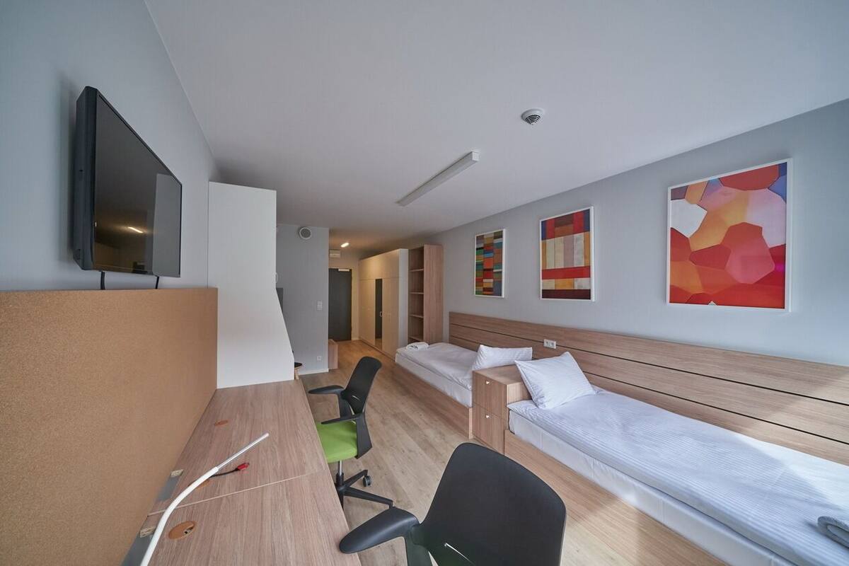 A hotel room with a desk, two single beds, a TV, and artwork on the walls.