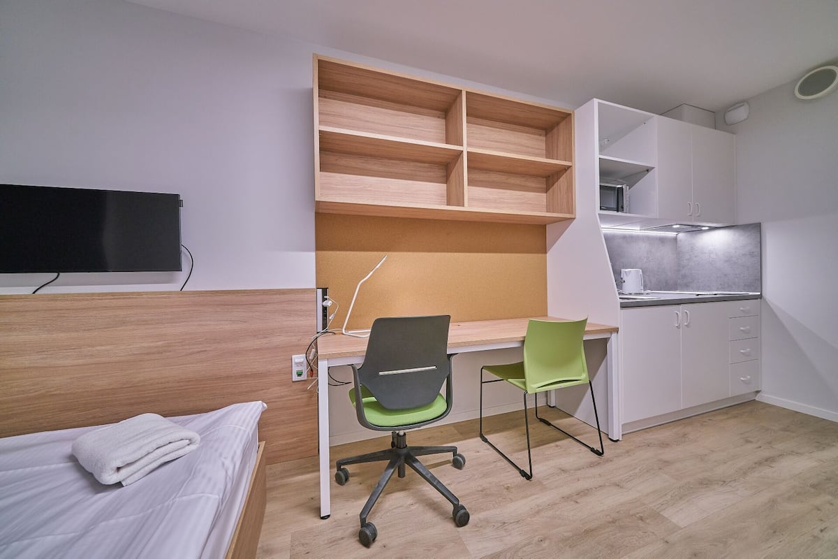 A compact room with a bed, a desk with a chair, a kitchenette, and a TV mounted on the wall.
