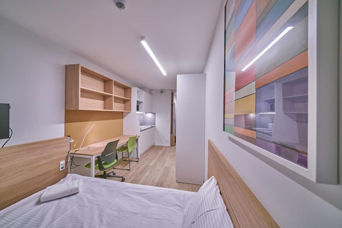 A compact hotel room with a bed, a desk, and a chair. There is a kitchenette in the background.