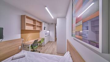 A compact hotel room with a bed, a desk, and a chair. There is a kitchenette in the background.