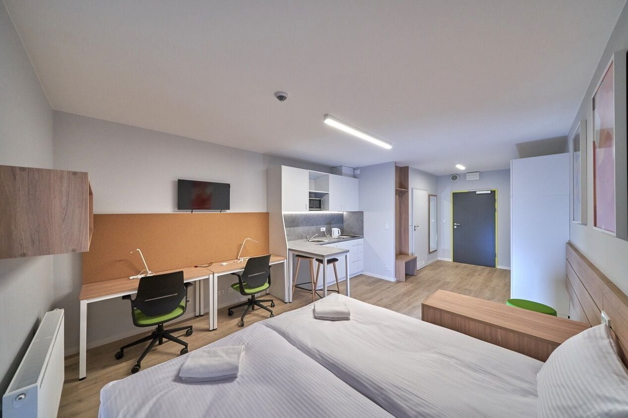 A hotel room with two single beds, a desk with a chair, a kitchenette, and a bathroom with a shower.