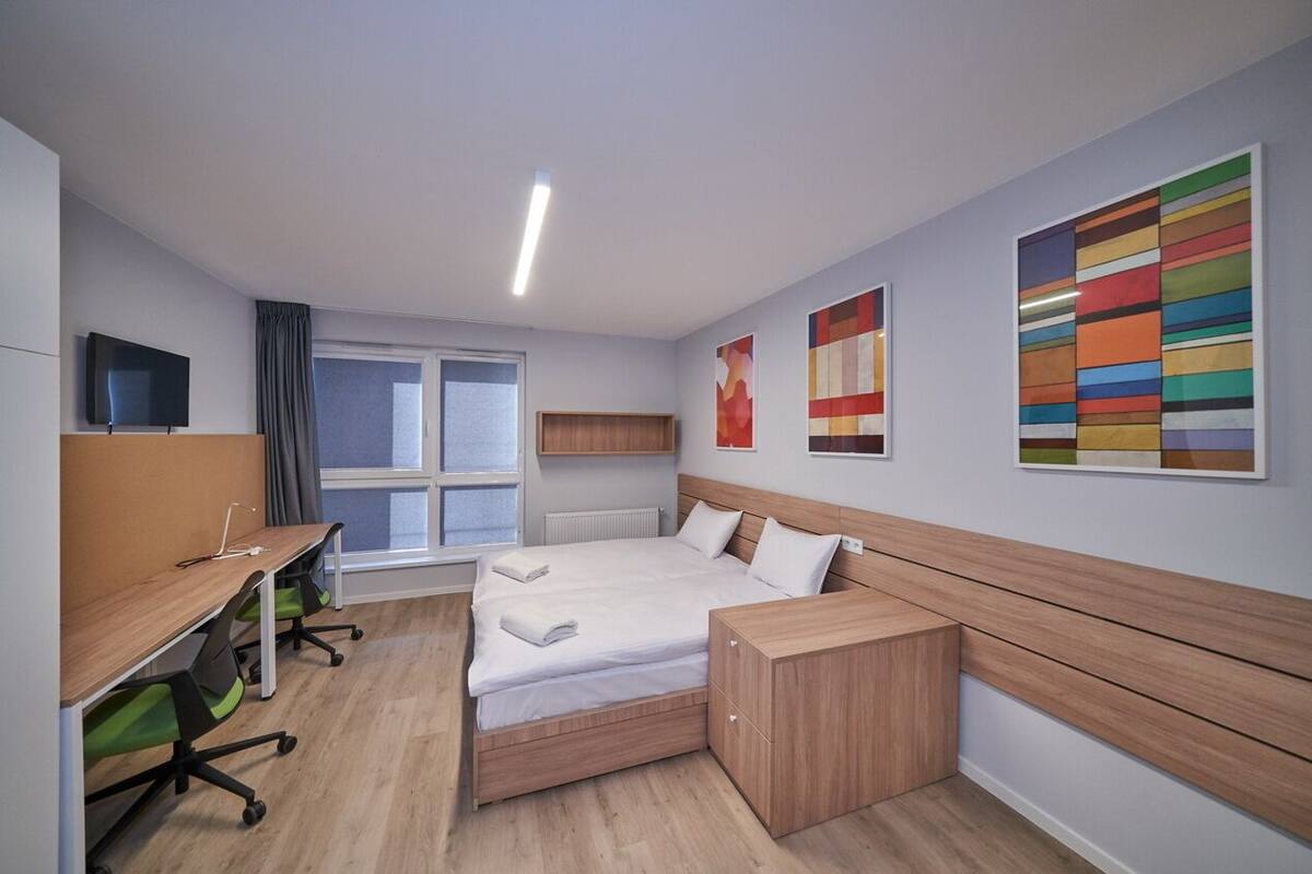 A modern hotel room with a bed, desk, and chair, a window with curtains, and colorful artwork on the walls.
