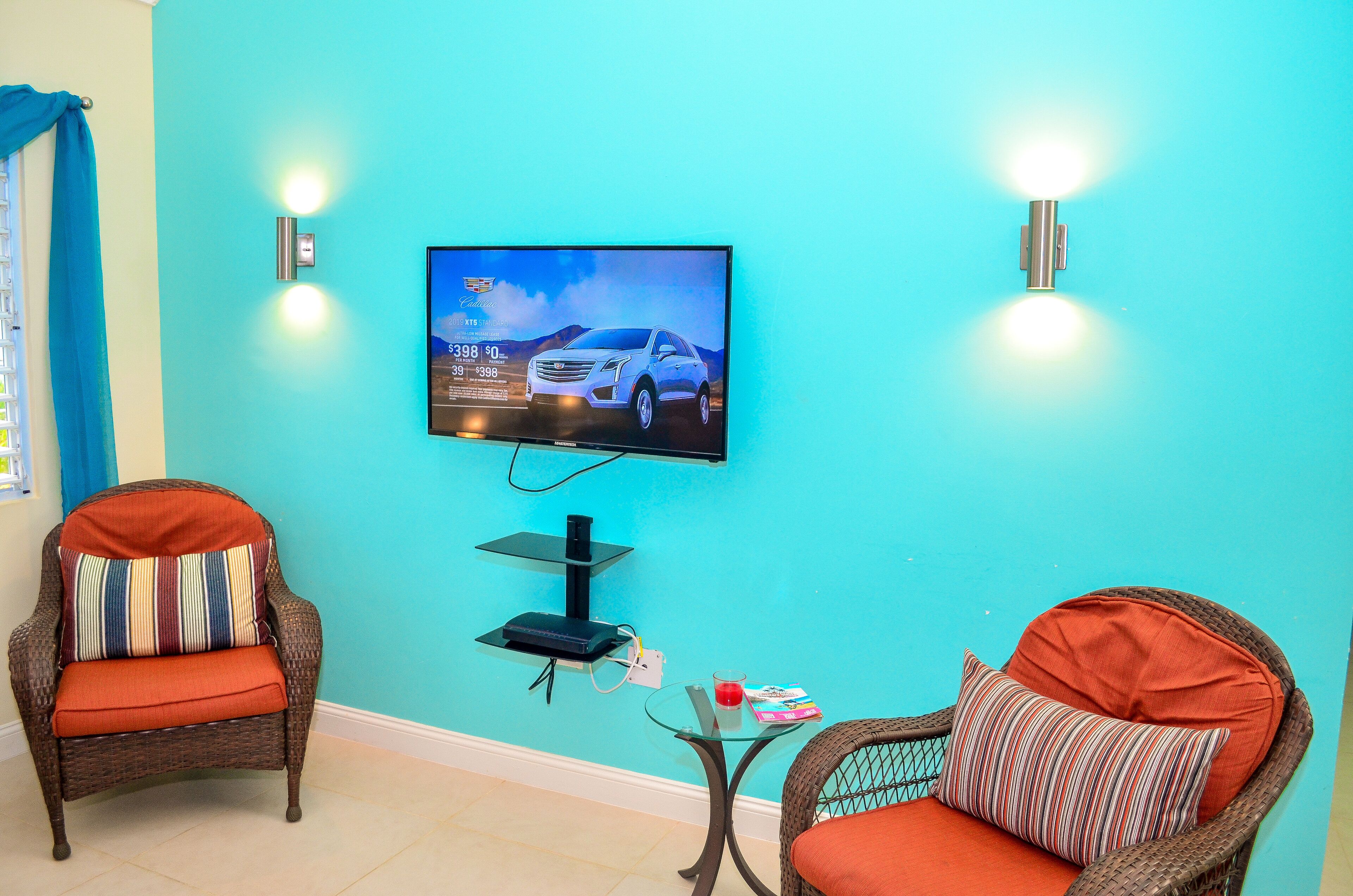 Een kamer met turquoise wanden, een wandgemonteerde flatscreen-tv waarop een auto reclame wordt weergegeven, twee rieten stoelen met oranje kussens, en een kleine glazen tafel met een afstandsbediening en een boek.