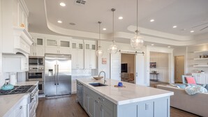 Une cuisine spacieuse avec des meubles blancs, un îlot central, des appareils en acier inoxydable et des luminaires suspendus.