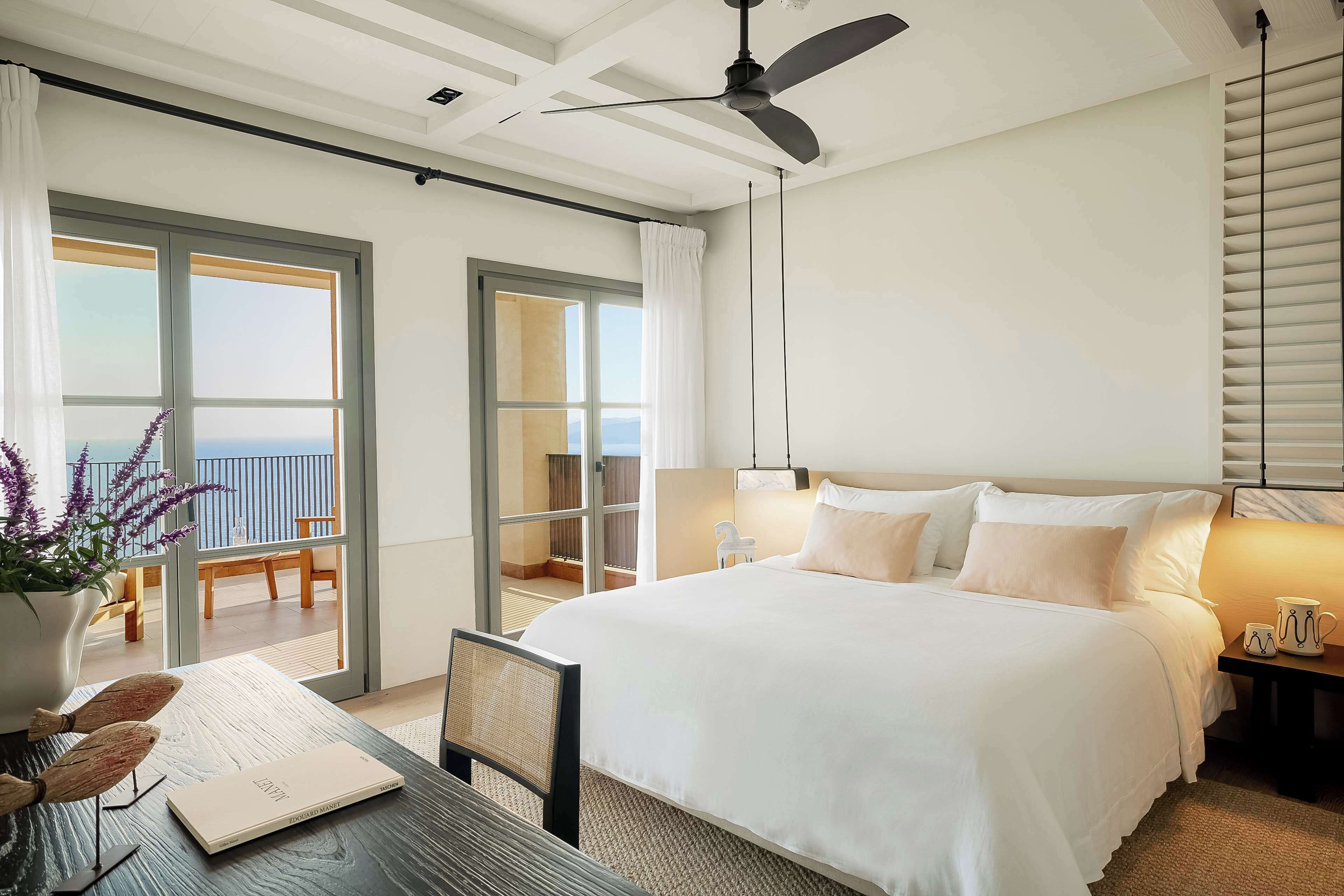 Une chambre d’hôtel avec un grand lit, une table à manger, un ventilateur de plafond et une vue sur la mer par les fenêtres.