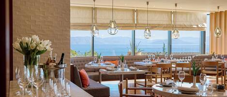 Un restaurante con vistas a las montañas, que cuenta con una zona de bar con copas de vino, un área de estar con mesas y sillas, y un amplio ventanal con cortinas.