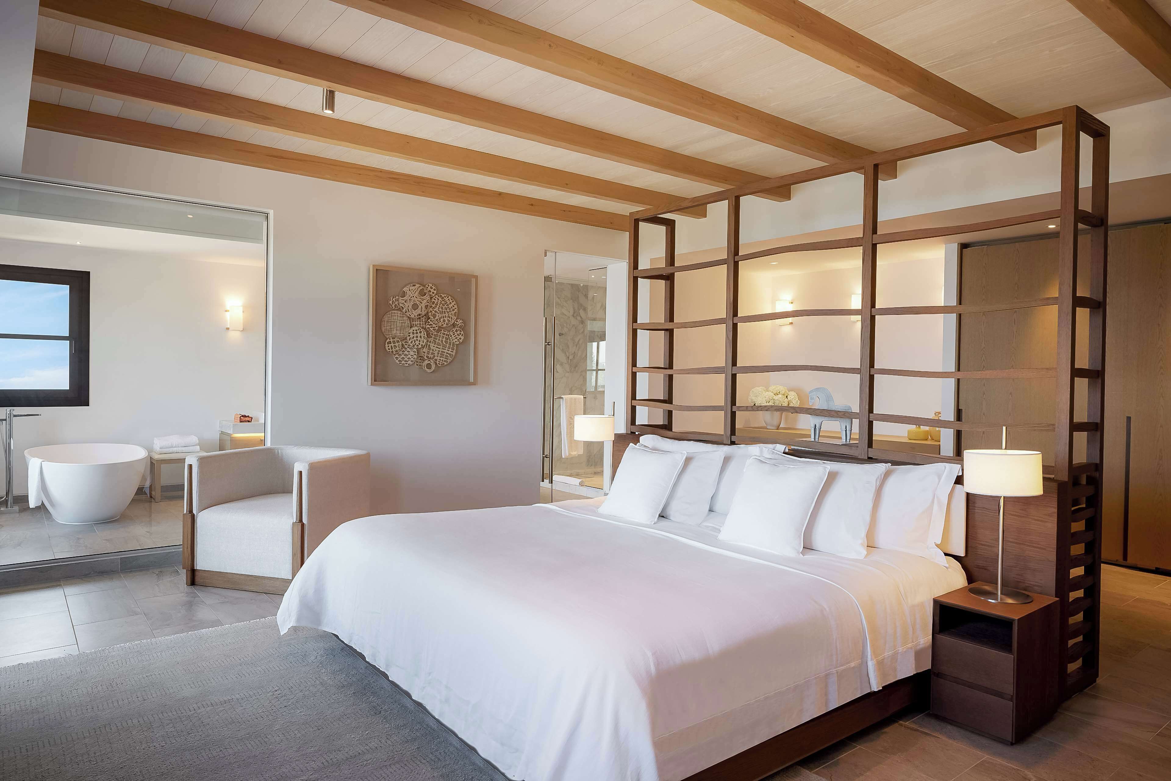 A modern hotel room with a large bed, a sitting area with a chair, a bathtub, and a wooden ceiling.