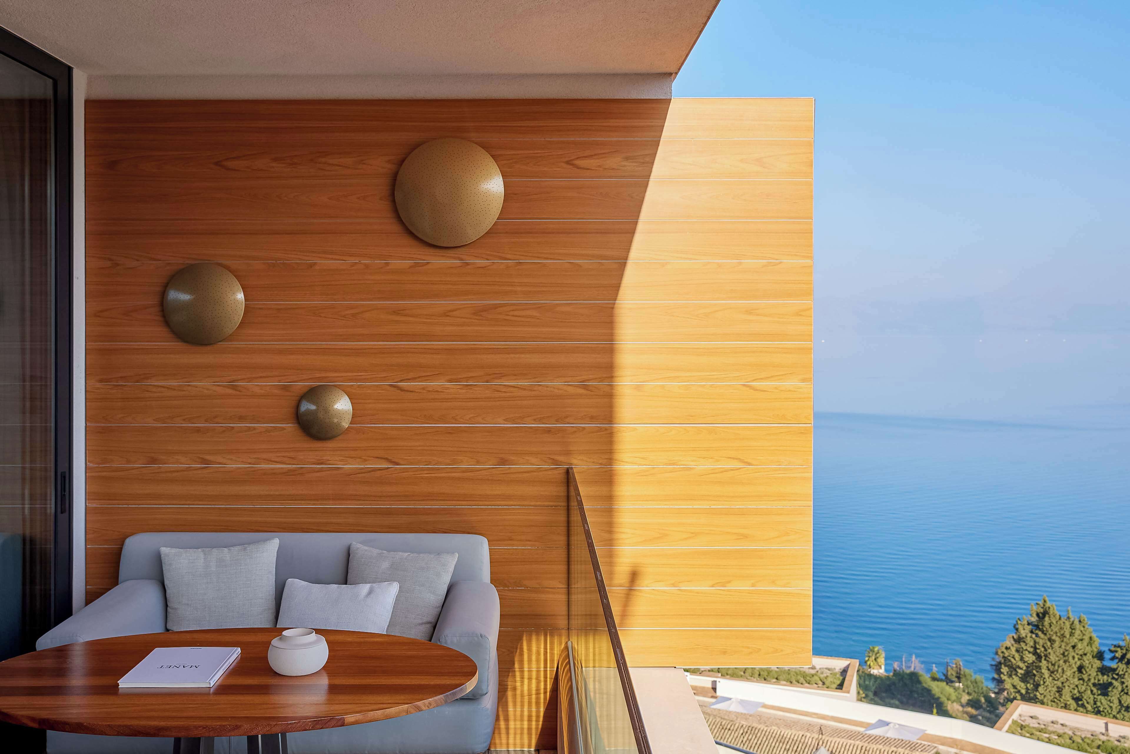 Quarto moderno com parede de madeira, uma mesa redonda, um sofá e vista para o mar.