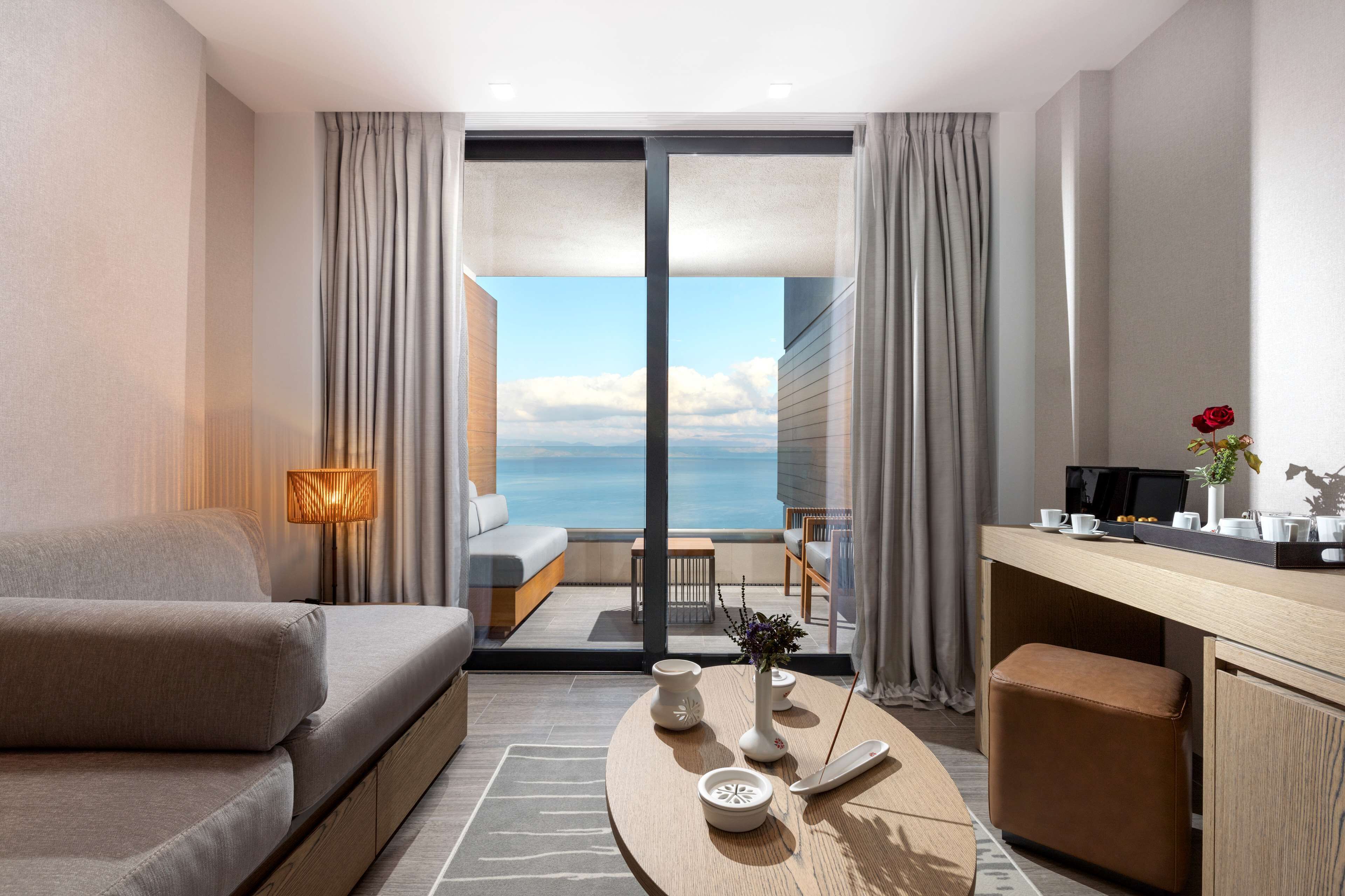 A modern hotel room with a sofa, a small table with a vase and a remote control, a desk with a computer and a vase with a rose, and a view of the sea through large windows.