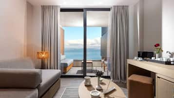Ein modernes Hotelzimmer mit einer Couch, einem kleinen Tisch mit einer Vase und einer Fernbedienung, einem Schreibtisch mit einem Computer und einer Vase mit einer Rose, sowie Meerblick durch große Fenster.