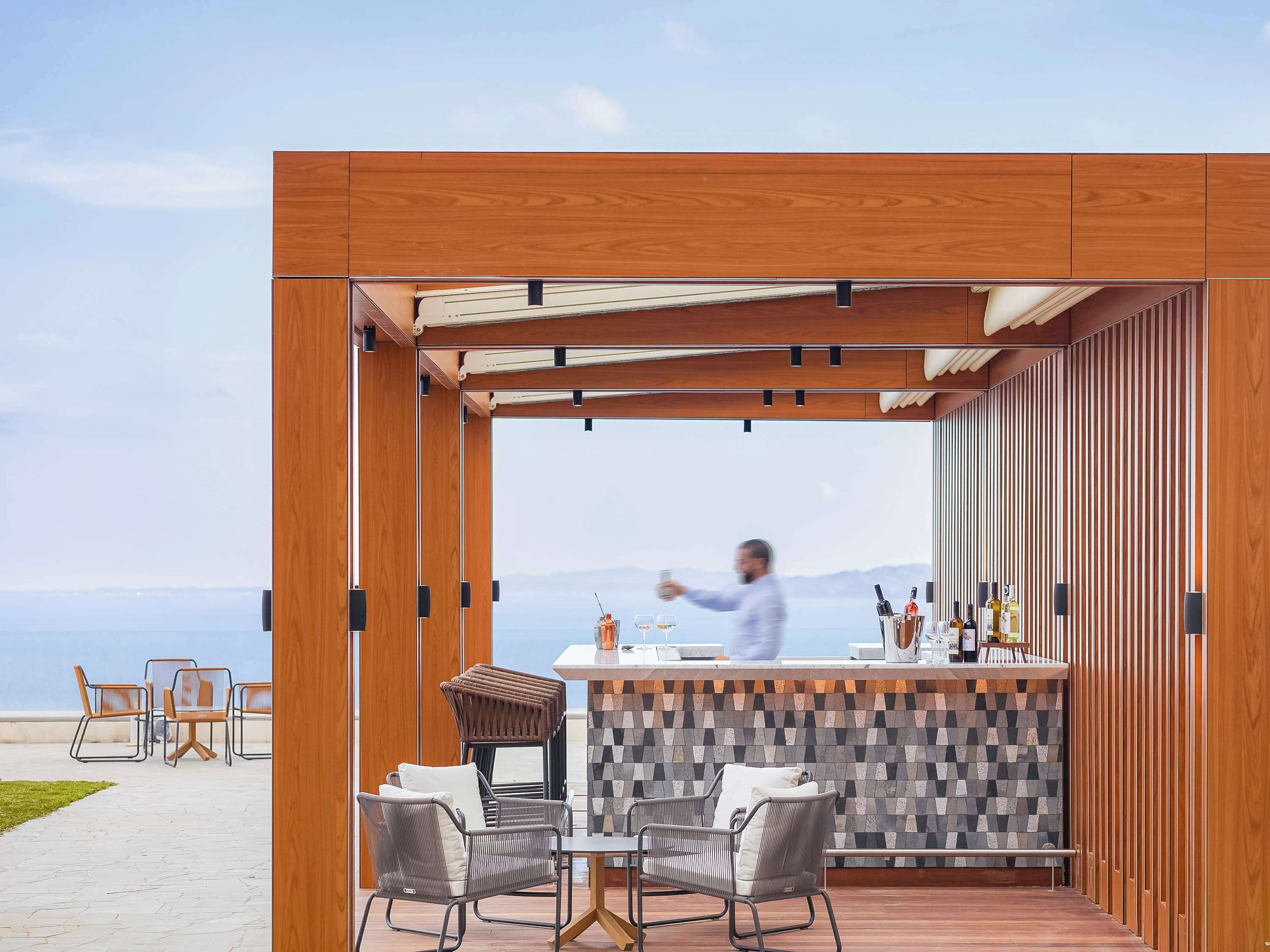 Un espace bar extérieur moderne avec vue sur la mer, comprenant une pergola en bois, des chaises en osier et un mur recouvert de carreaux.