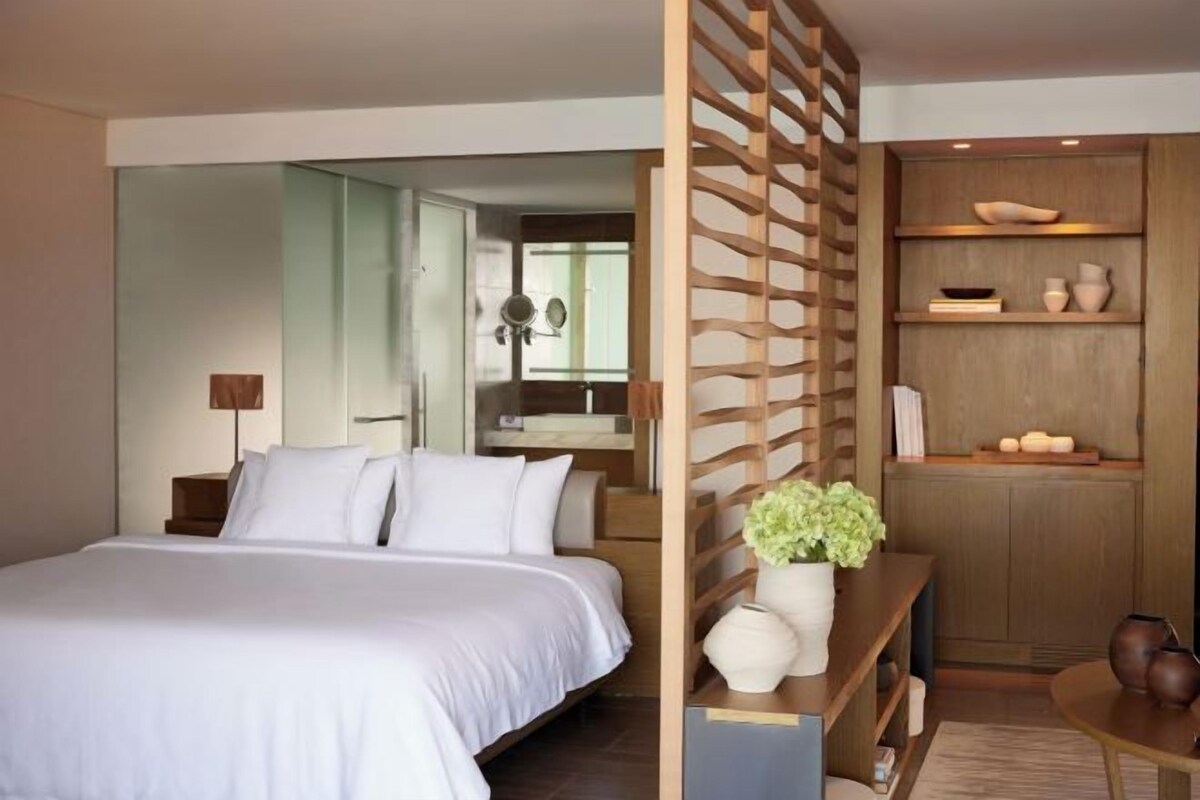 Habitación de hotel moderna con una cama grande, un separador de madera, una mesita con un florero y estantes con objetos decorativos.