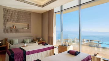 A hotel room with two beds, a large window with a view of the sea, a decorative wall, and a small table with a vase.