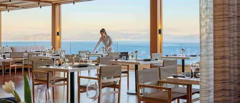 A restaurant with tables set for dining, a waiter attending to guests, and a view of the sea.