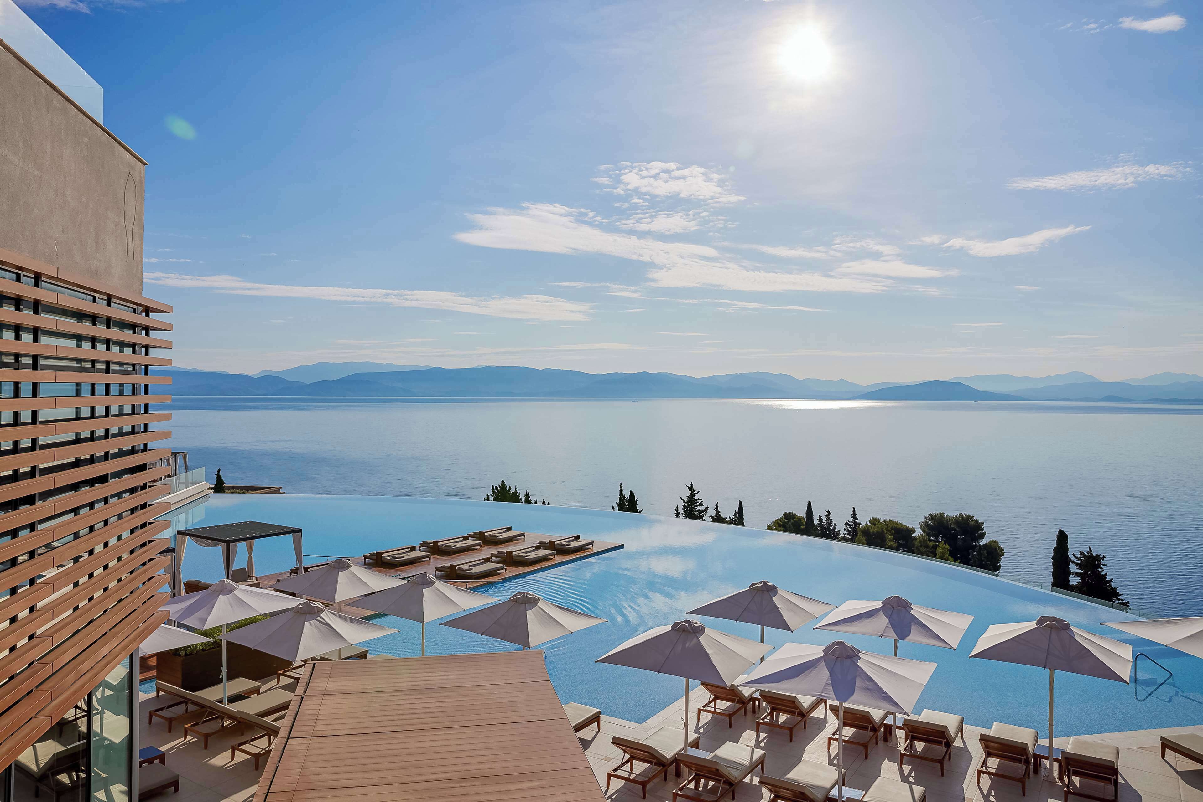 A rooftop infinity pool with lounge chairs and umbrellas, overlooking a calm lake and distant mountains.