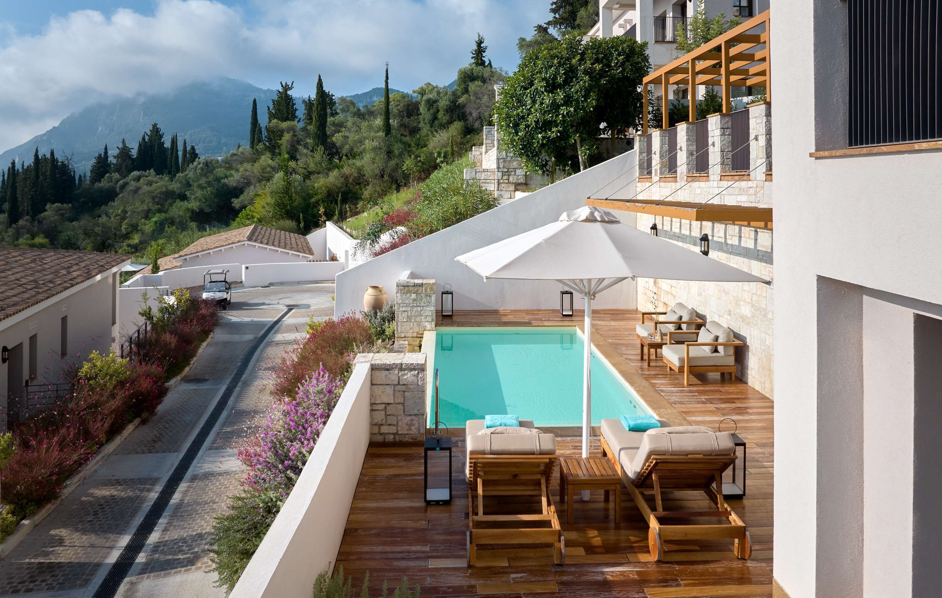 A rooftop pool area with wooden loungers, a white umbrella, and a view of a mountainous landscape.