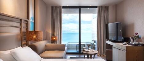 A modern hotel room with a bed, a sofa, a desk, a television, and a view of the sea.