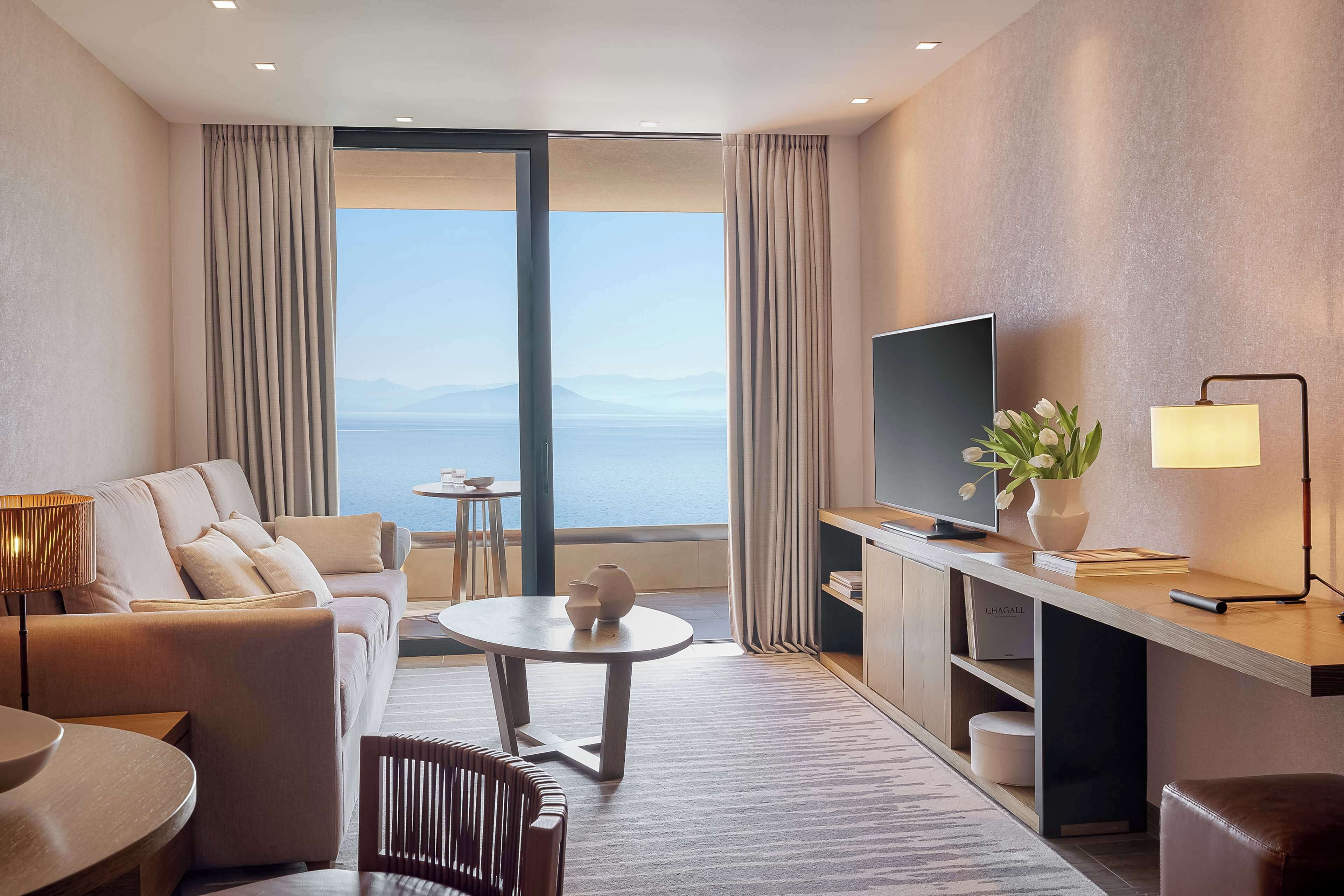 A modern hotel room with a large window, a sofa, a dining table, a TV, and a desk with a lamp.