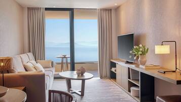 Ein modernes Hotelzimmer mit großem Fenster, einer Couch, einem Esstisch, einem Fernseher und einem Schreibtisch mit Lampe.