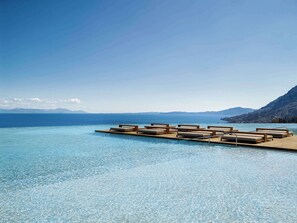 Vista despejada de una piscina con sillones de descanso y una terraza de madera que se extiende sobre el agua, rodeada de montañas y un cielo claro.